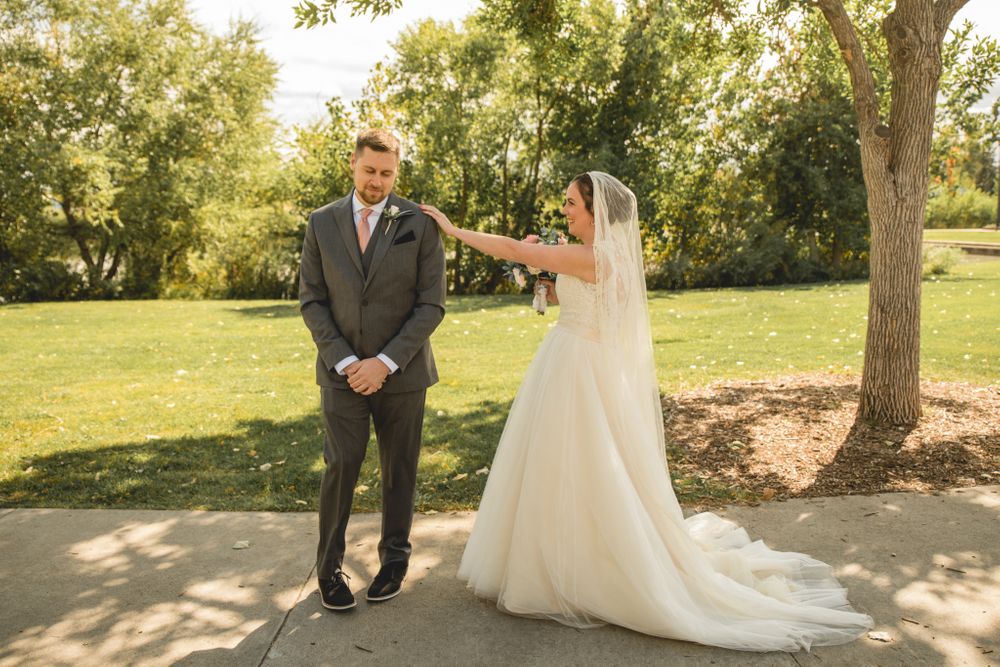 A bride taps her groom's shoulder during their first look moment in a sunny garden setting.