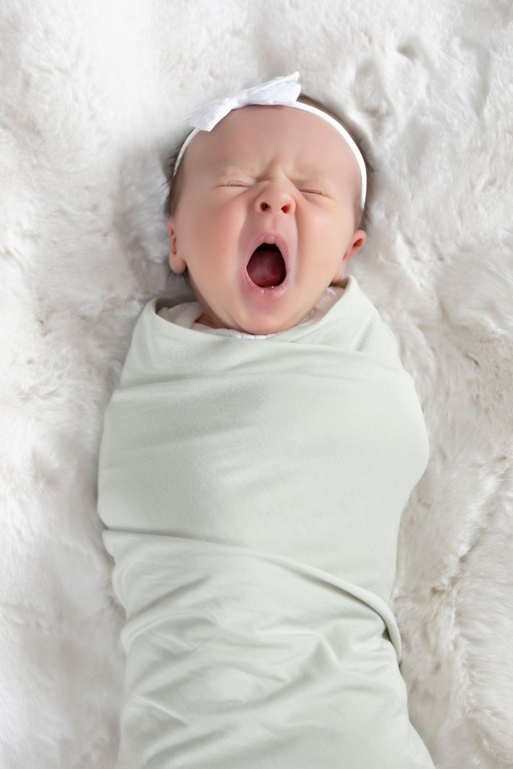 newborn baby swaddled in green blanket, white headband, she is yawning, eyes closed