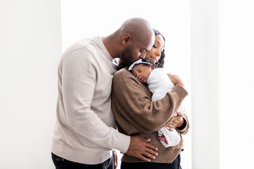 Man and woman stood, woman holding baby, head on her shoulders, Man is behind woman, kissing baby's head, close together
