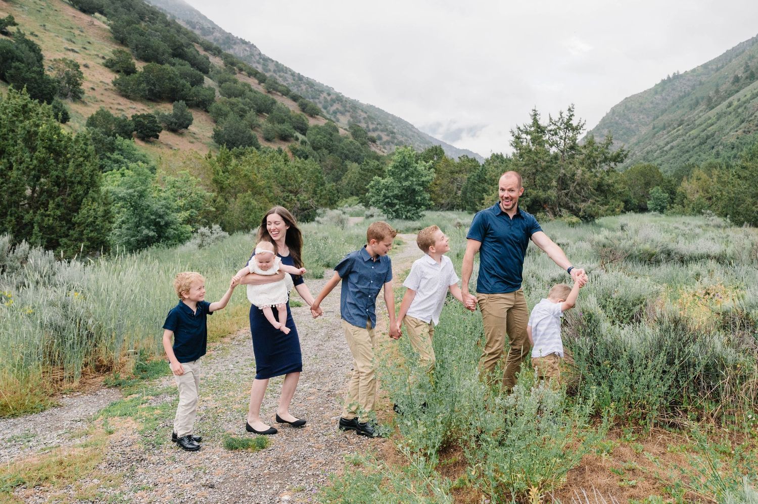 Family walks together holding hands along mountain trail surrounded by sage brush and hills.