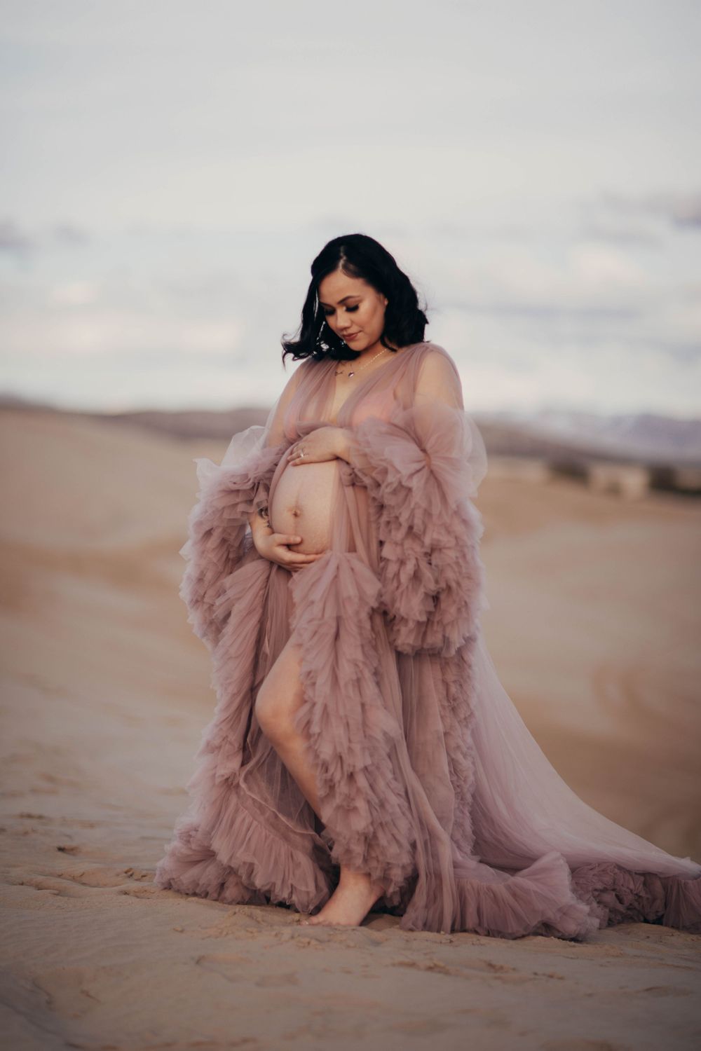 A pregnant woman in a flowing mauve tulle gown poses gracefully on sandy desert dunes.