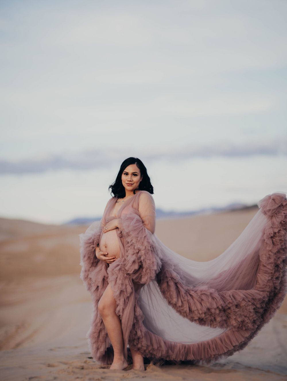 A pregnant woman in a billowing mauve tulle maternity gown poses on open sandy desert dunes.