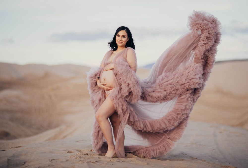 Pregnant woman in flowing pink tulle gown poses gracefully on desert sand dunes at sunset.