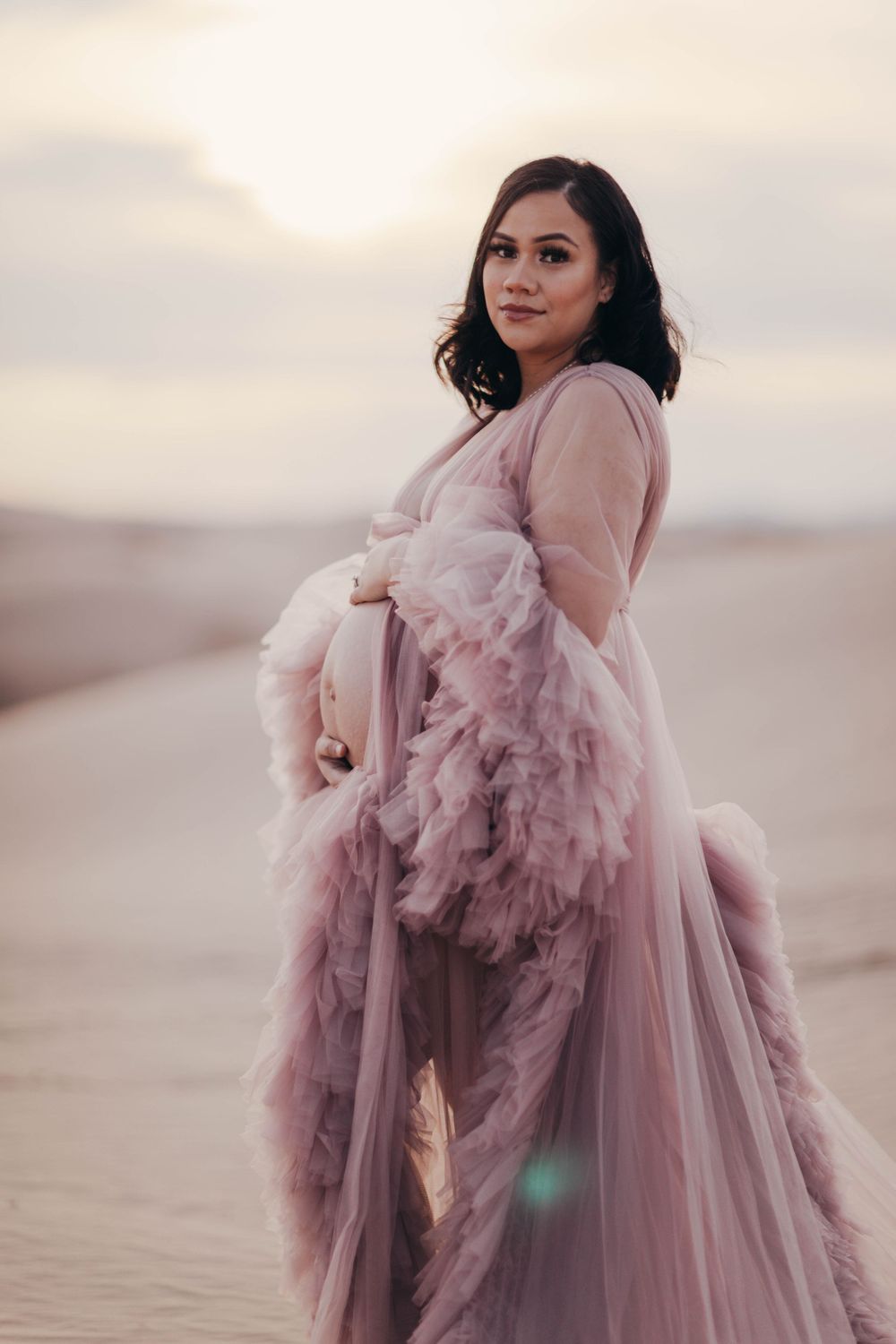Expectant mother in mauve ruffled tulle maternity gown stands serenely on sandy desert dunes.