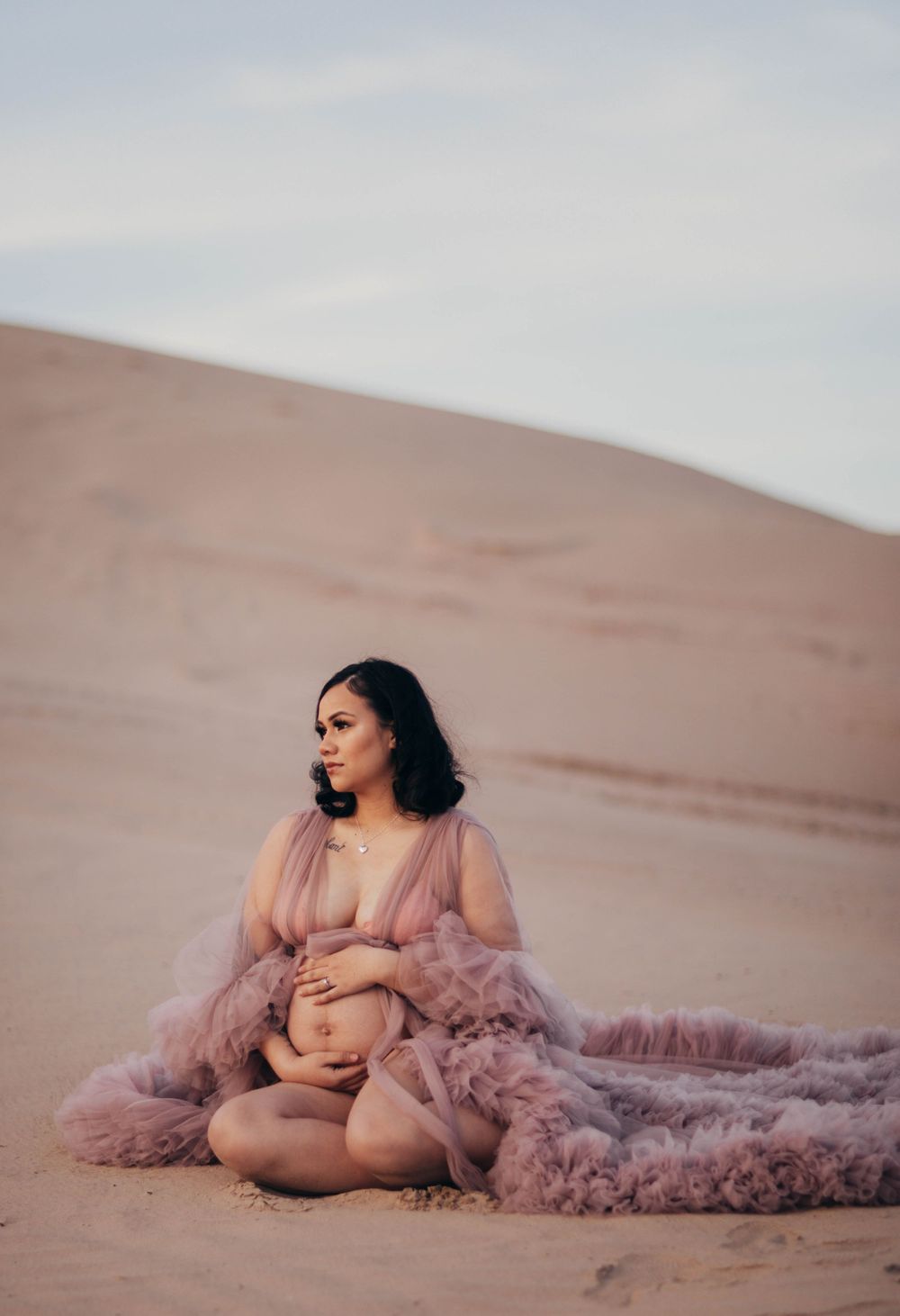 Pregnant woman in sheer pink tulle gown sits elegantly on golden sand dunes at dusk.