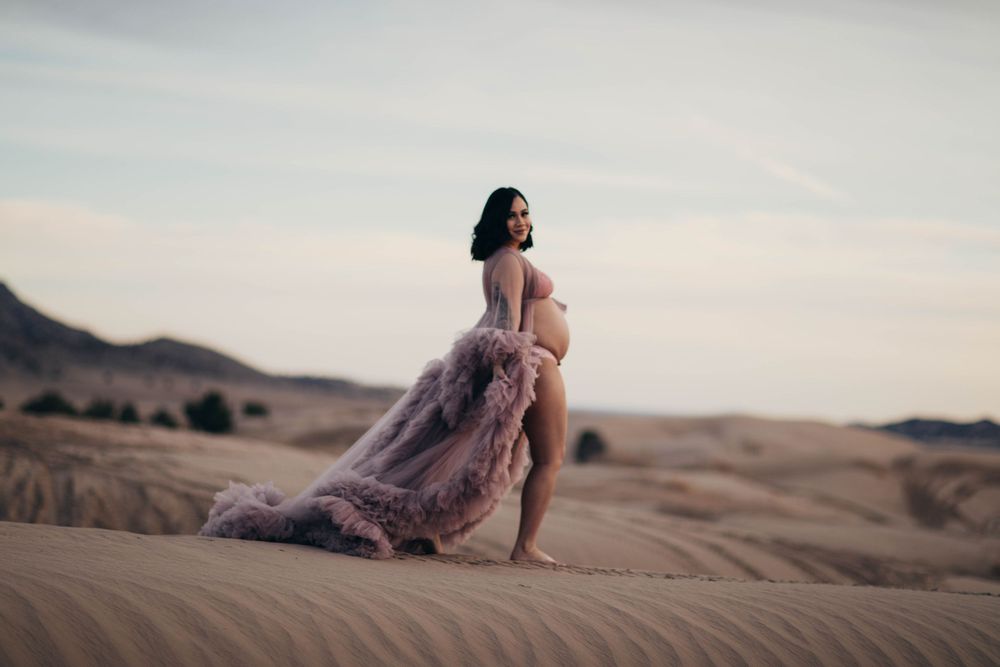 Barefoot expectant mother in flowing pink ruffled gown walks across vast desert sand dunes.