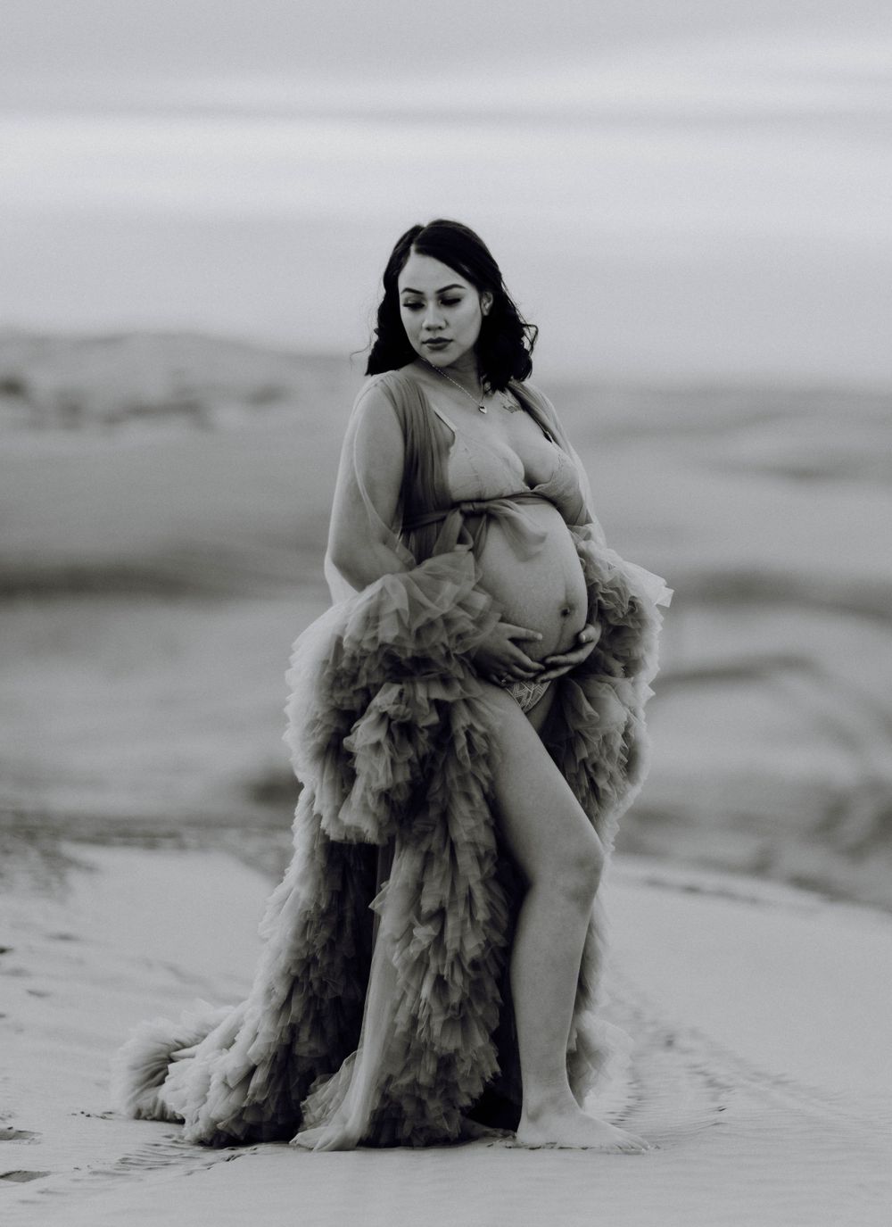 Black and white maternity photo of pregnant woman in ruffled tulle gown on sand dunes.