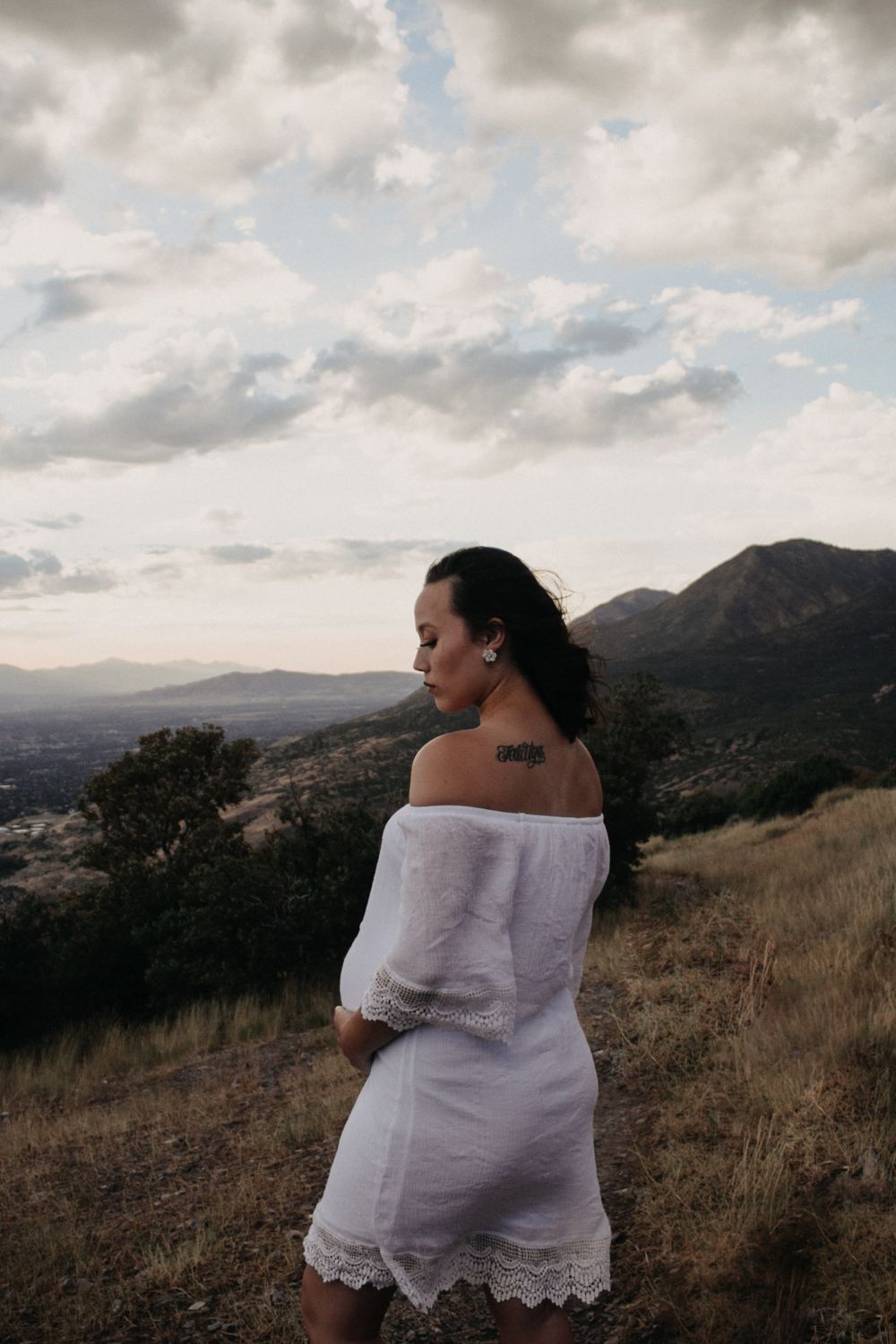 A pregnant woman in a white off-shoulder dress stands on a hillside with mountain views behind her.