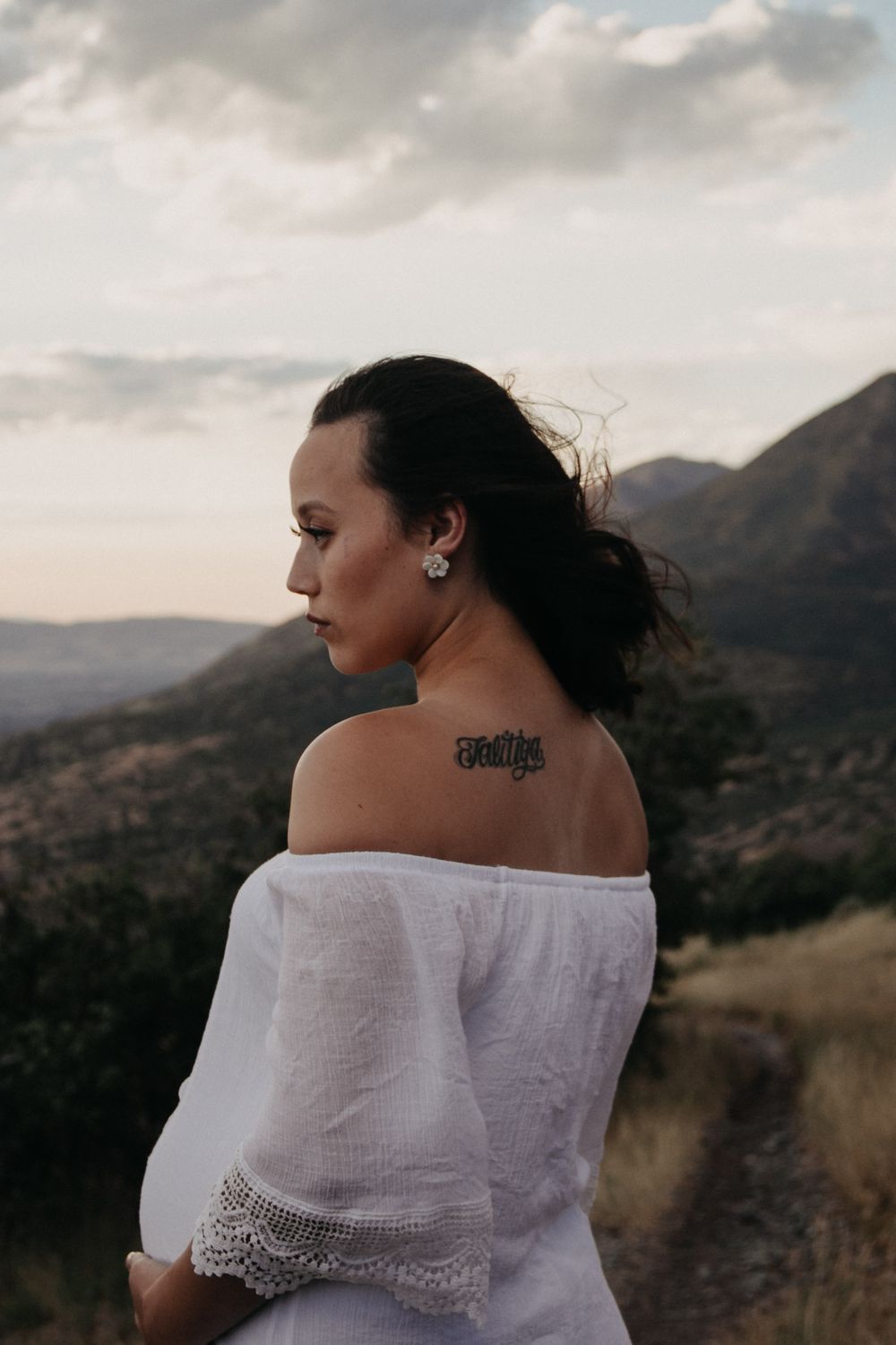 Pregnant woman in white off-shoulder dress showing back tattoo with mountain landscape backdrop.