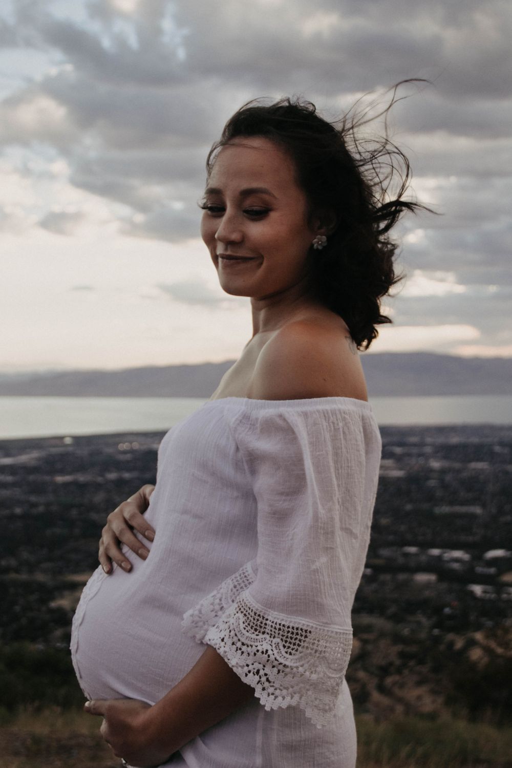 Smiling pregnant woman in white lace off-shoulder dress cradling belly outdoors at sunset.