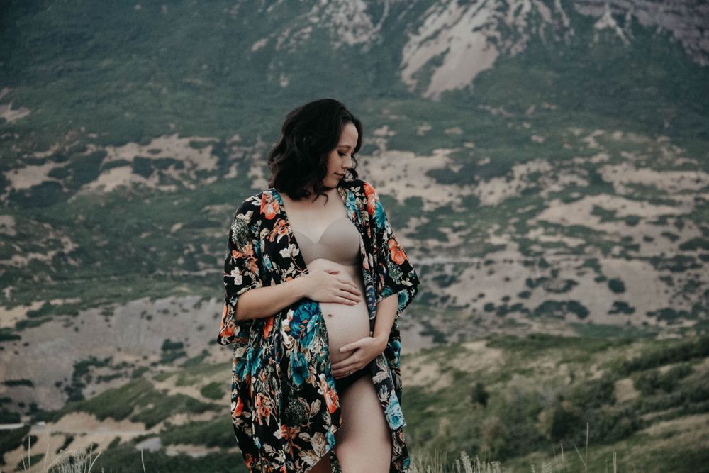 Pregnant woman in floral kimono and nude bodysuit cradling belly against mountain landscape.
