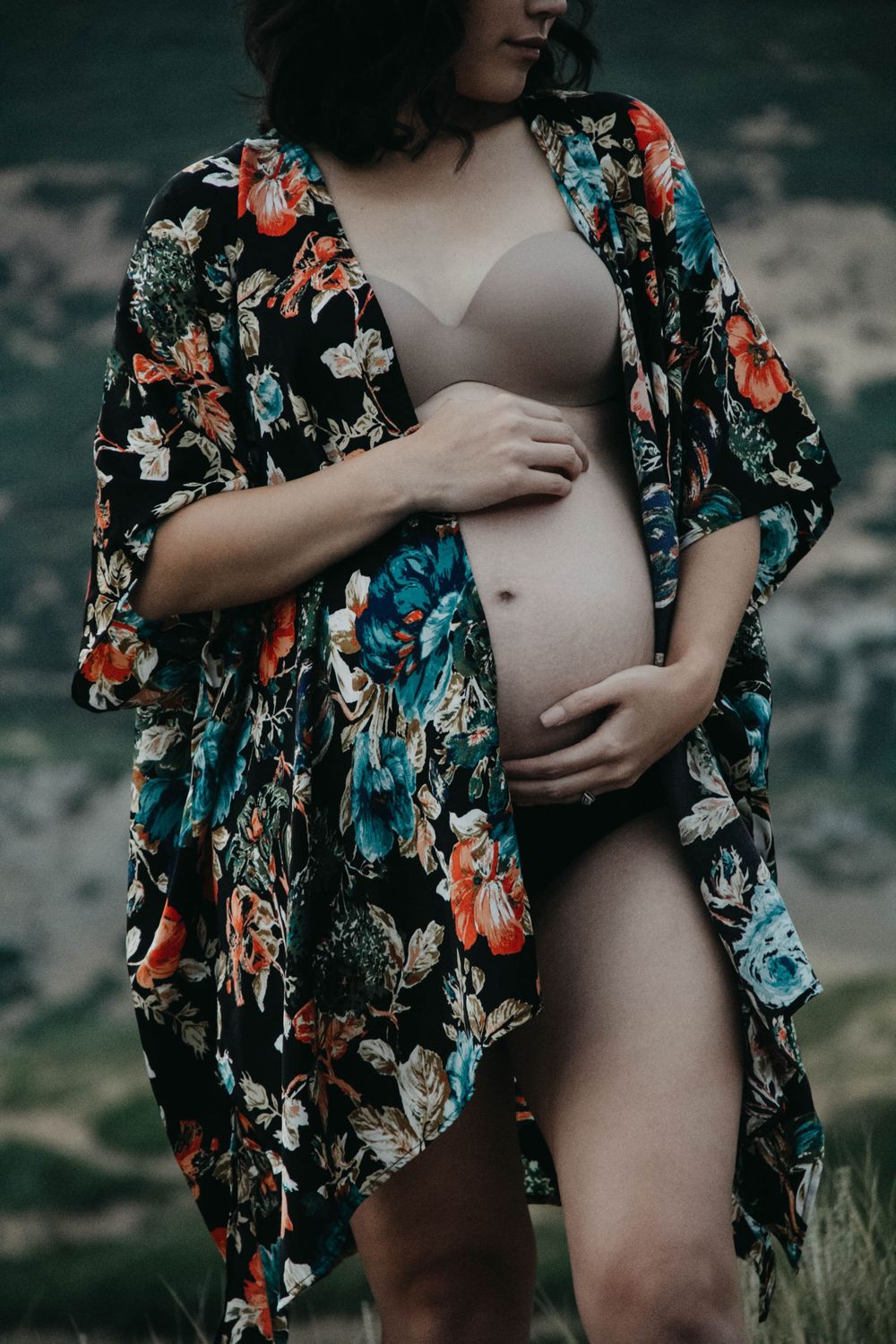 Pregnant woman in colorful floral kimono and nude bodysuit showcasing baby bump outdoors.