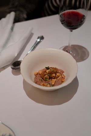Small white bowl containing nuts and garnish beside a glass of red wine.