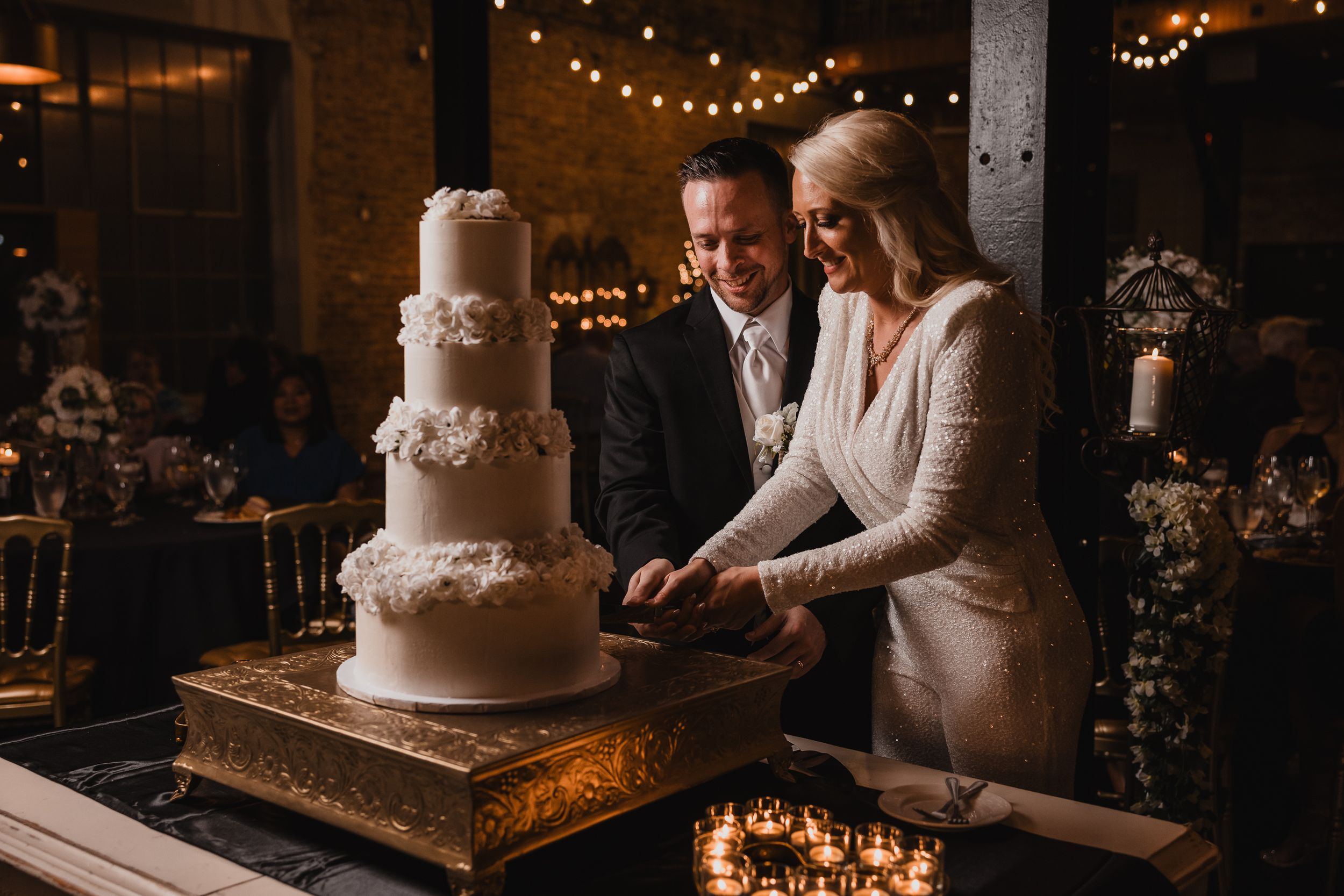 Elegant white wedding cake displayed at a rustic venue with warm string lights creating a romantic ambiance.