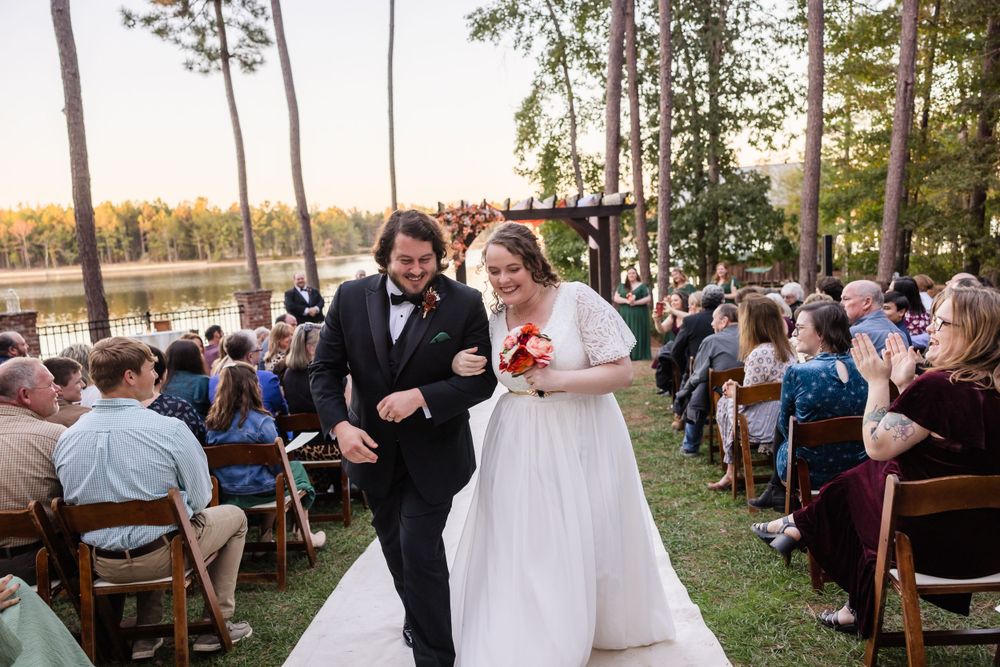 Outdoor wedding reception by a lake with wooden benches and guests gathered at sunset.