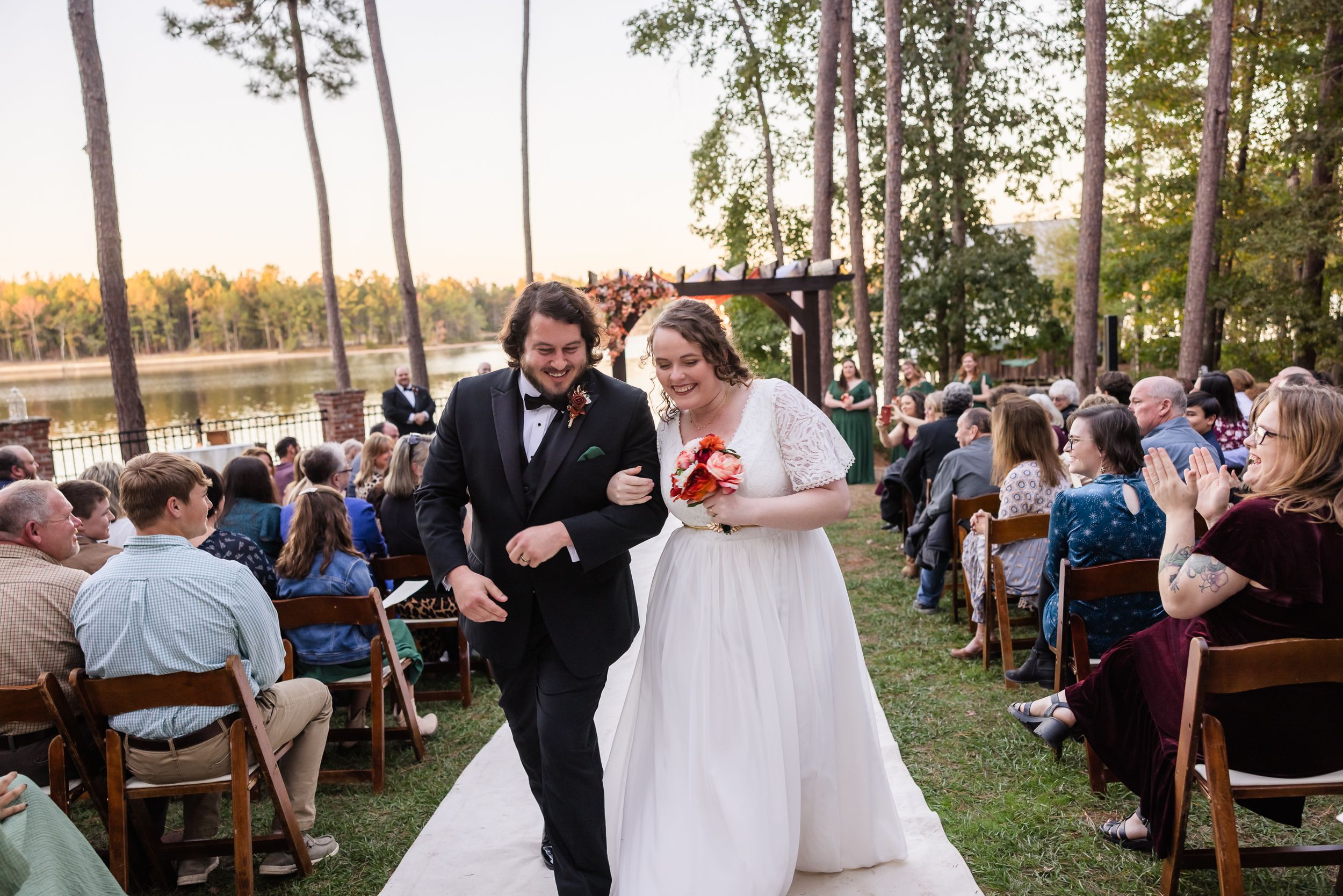 Outdoor wedding reception by a lake with wooden benches and guests gathered at sunset.