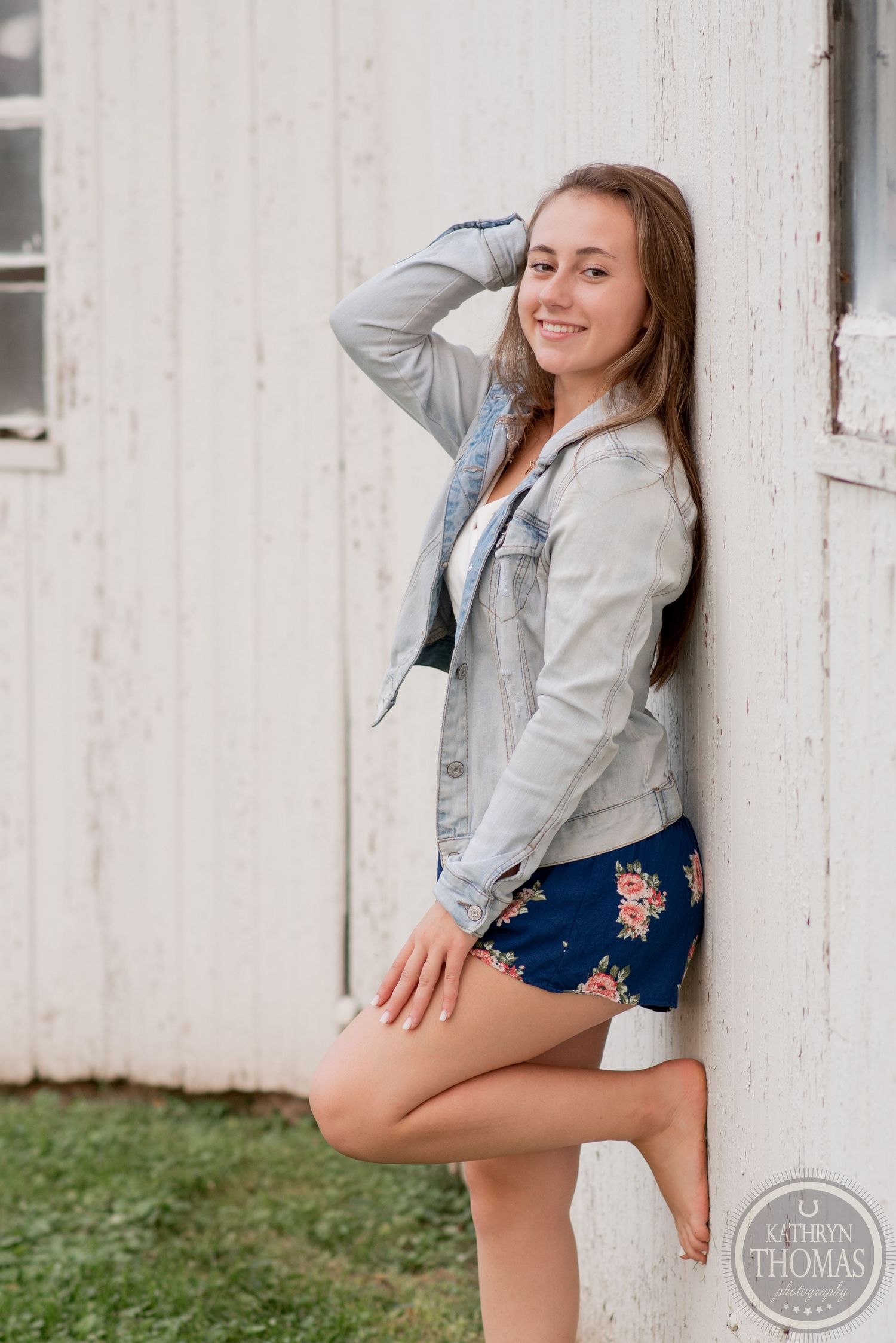 Casual portrait against white barn wall wearing light denim jacket and navy floral shorts.