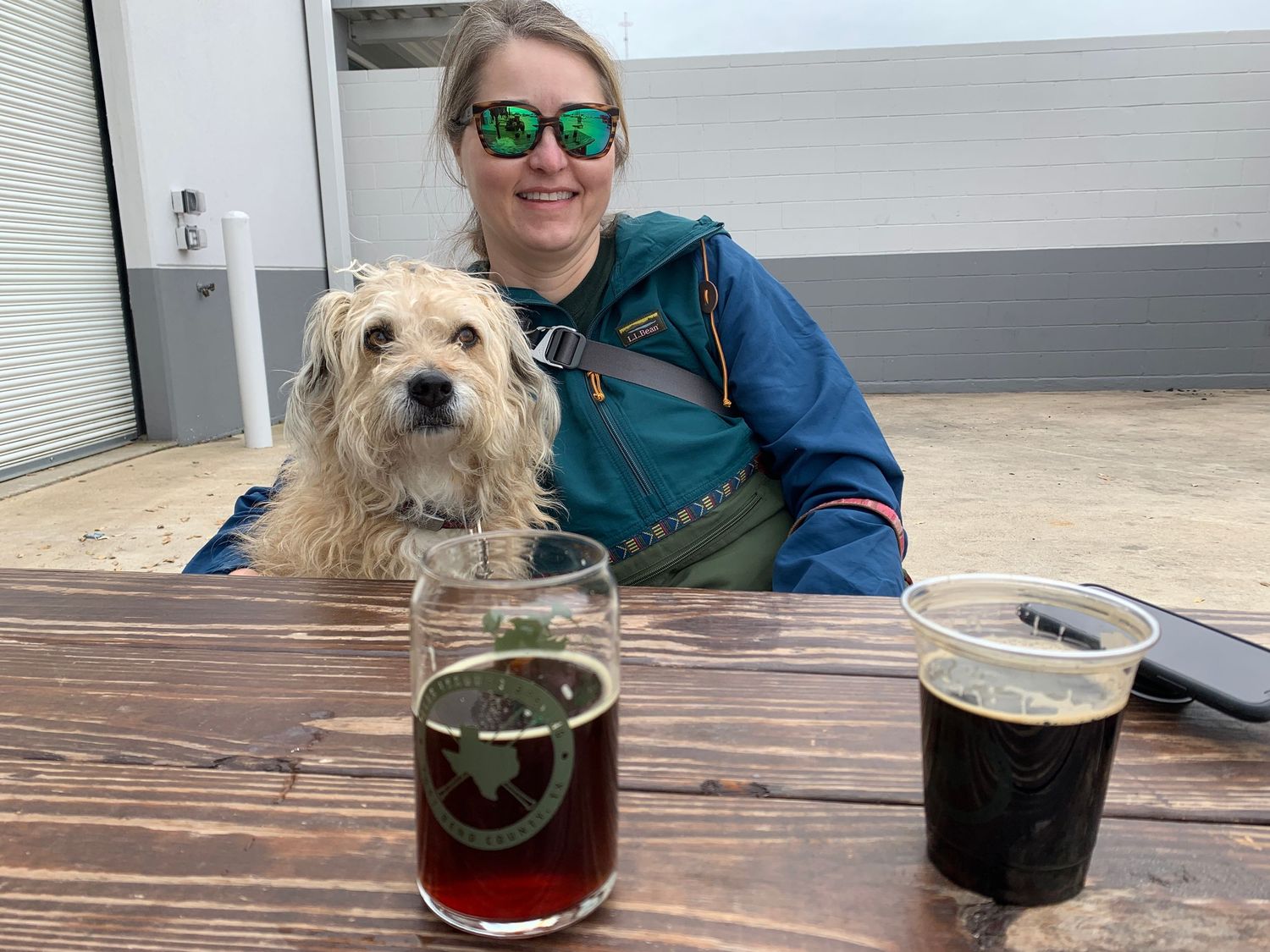 Shaggy dog sitting at outdoor brewery table with two dark beers.
