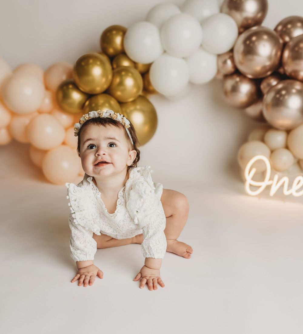 Baby in white dress crawling under balloon arch with gold and rose gold decorations.