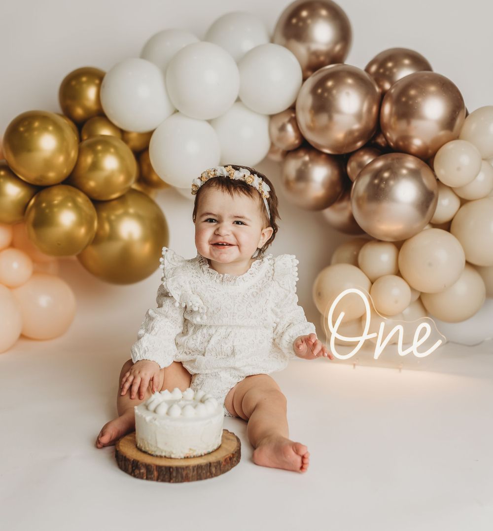 First birthday celebration with baby in white lace dress by balloon arch and smash cake.
