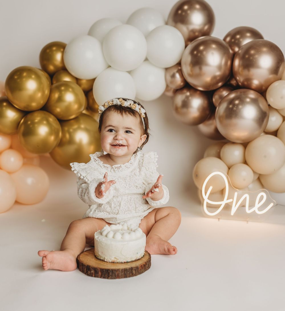 Smiling baby in white lace outfit sitting with birthday cake under metallic balloon arch.