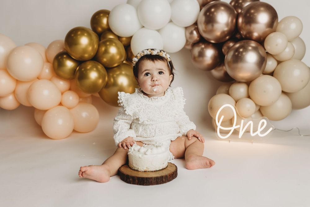 Baby wearing white lace outfit sitting with cake under balloon arch for first birthday shoot.