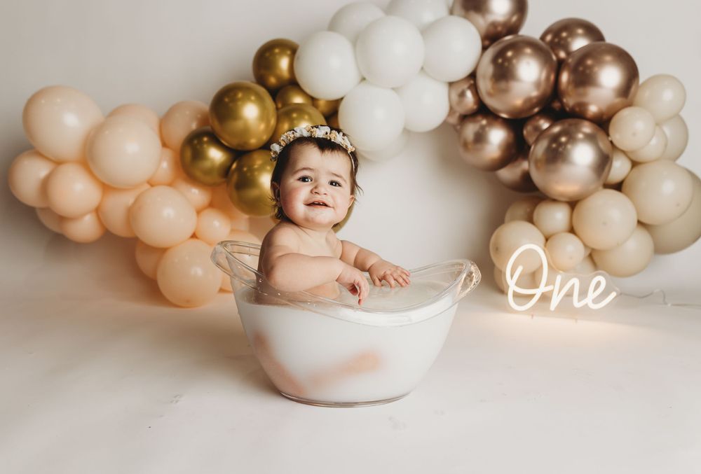 Baby sitting in white ceramic bowl under balloon arch during first birthday photoshoot.