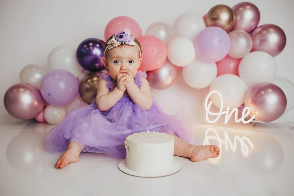 First birthday photo shoot setup with purple tulle dress, white cake, and pink and purple balloon arch backdrop.