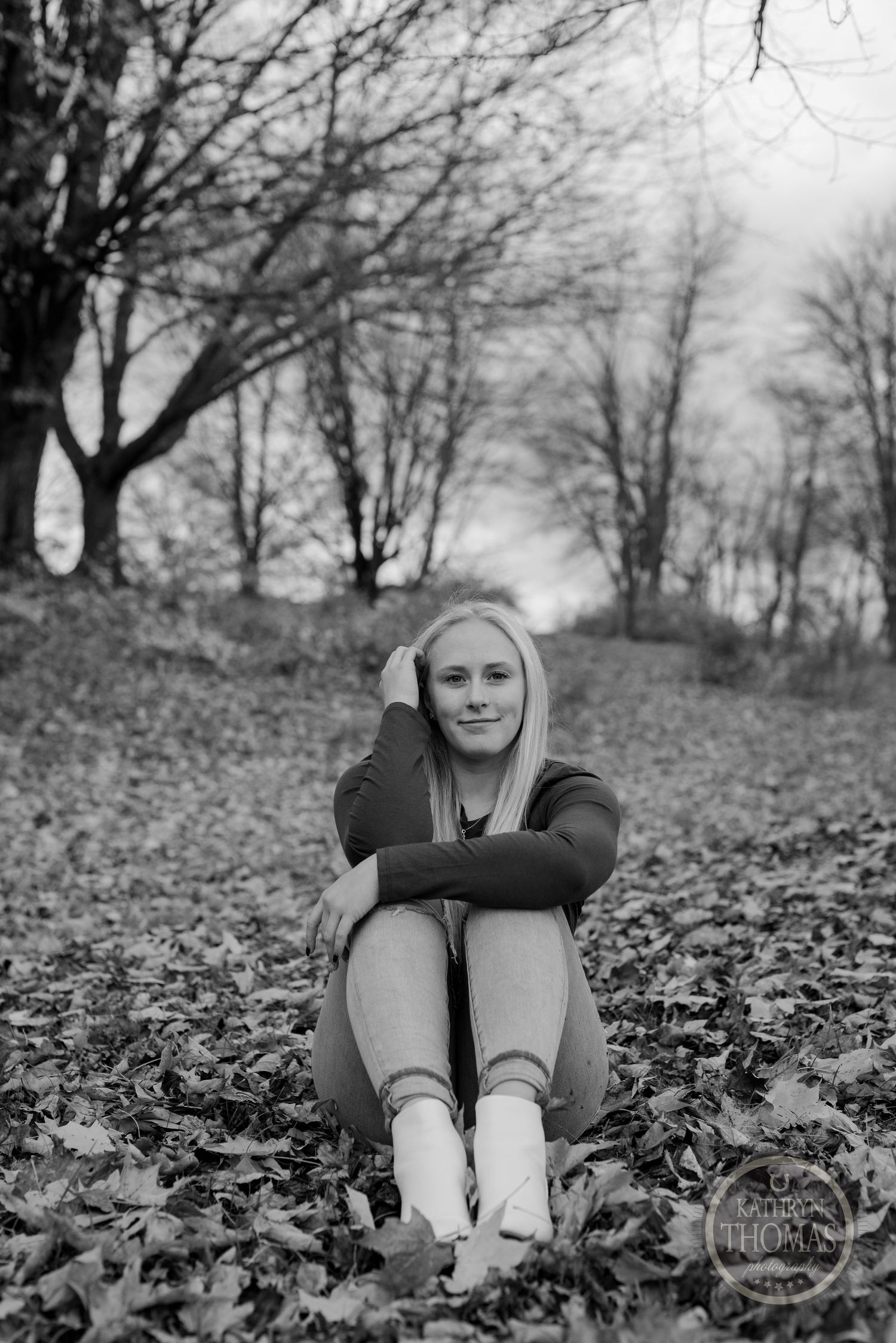 Person sitting on fallen leaves wearing dark top and light pants in monochrome woodland scene.