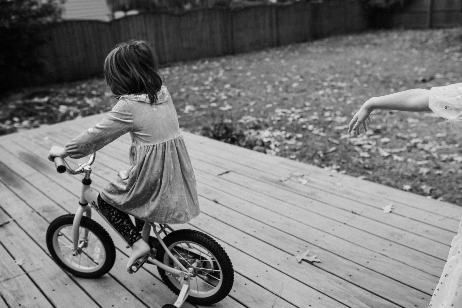girl riding bike on porch with another child's hand on the right side of the frame