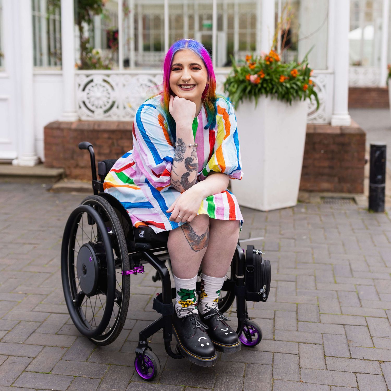 London rainbow haired disabled influencer posing sat smiling in their wheelchair wearing a rainbow outfit.