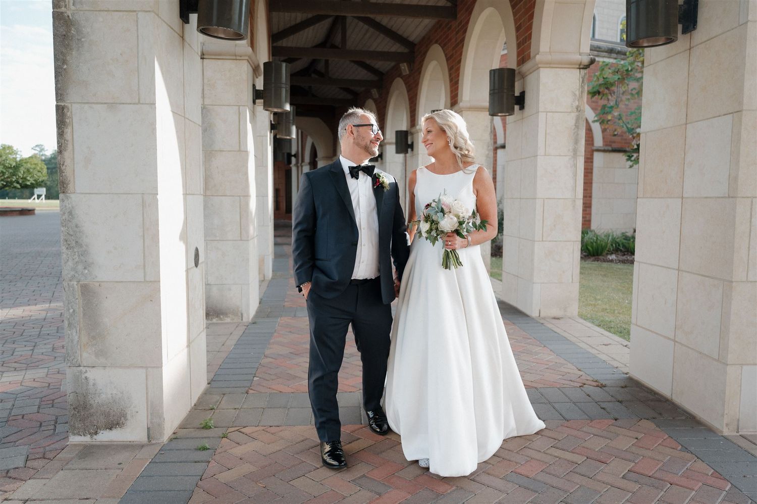 Couple strolling through elegant arched walkway with bouquet.