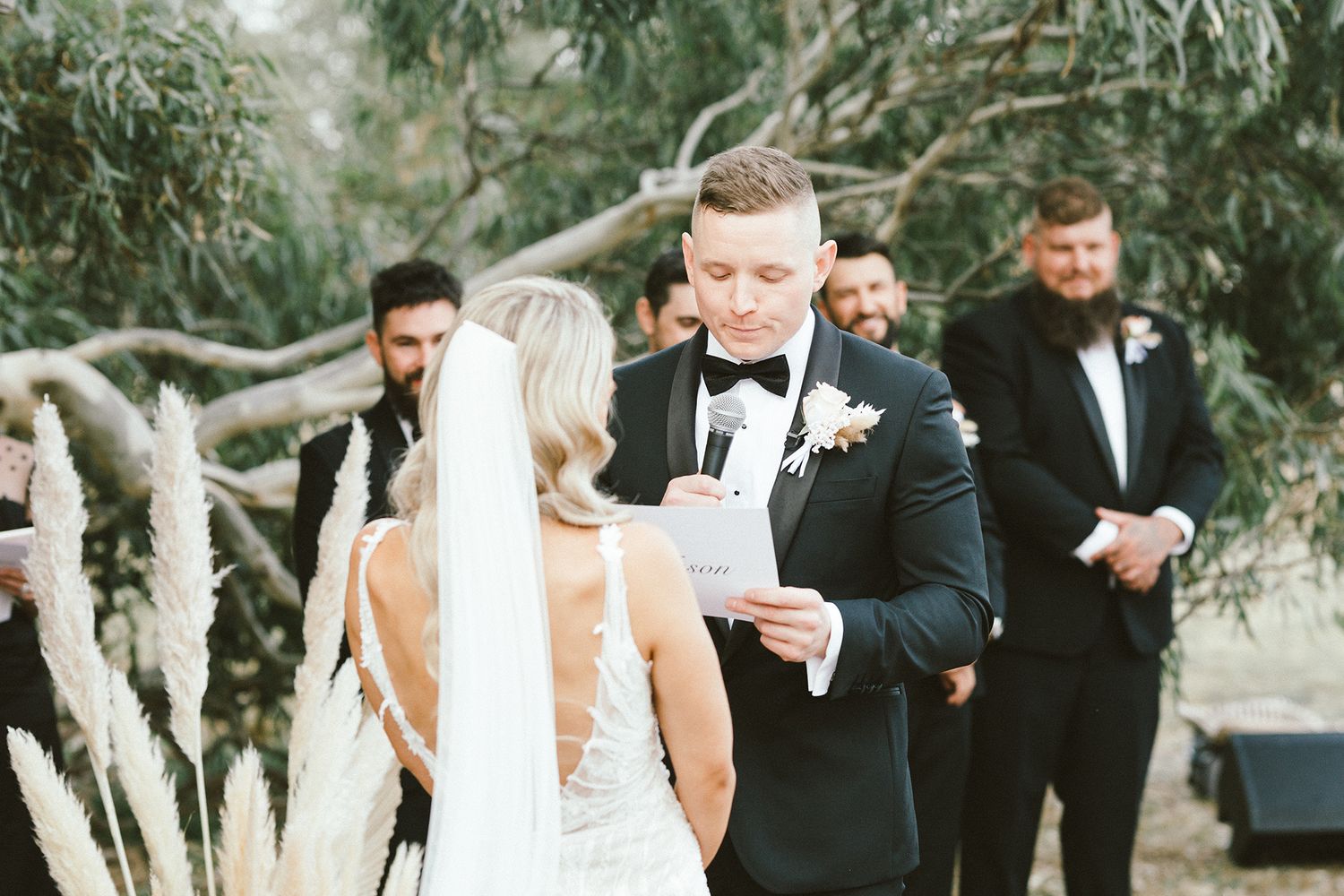 Wedding party members stand together during an outdoor ceremony beneath eucalyptus trees.
