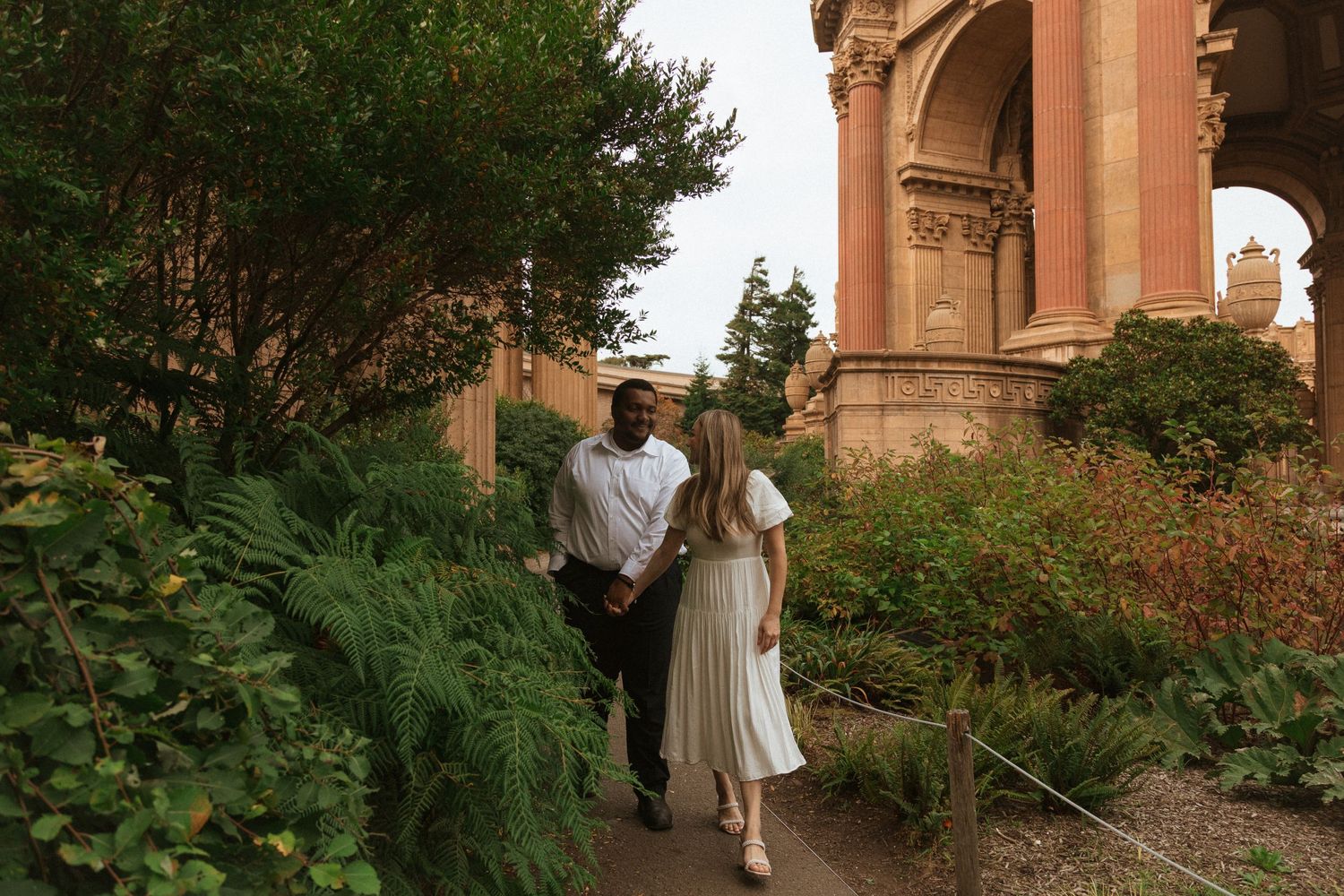A couple walking along a garden path beside an ornate stone archway and lush greenery.