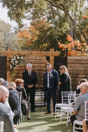 Wedding guests gather outdoors near a rustic wooden fence during an autumn ceremony with white chairs.
