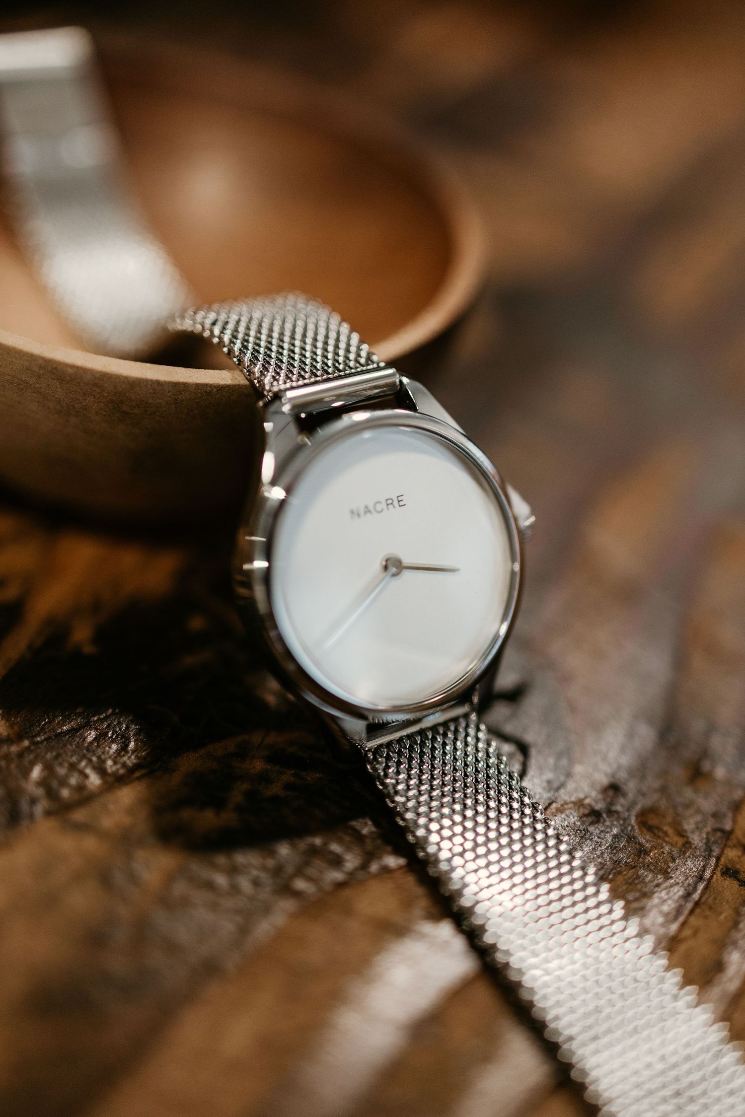 Modern minimalist watch with silver mesh strap captured in warm lighting on wooden surface.
