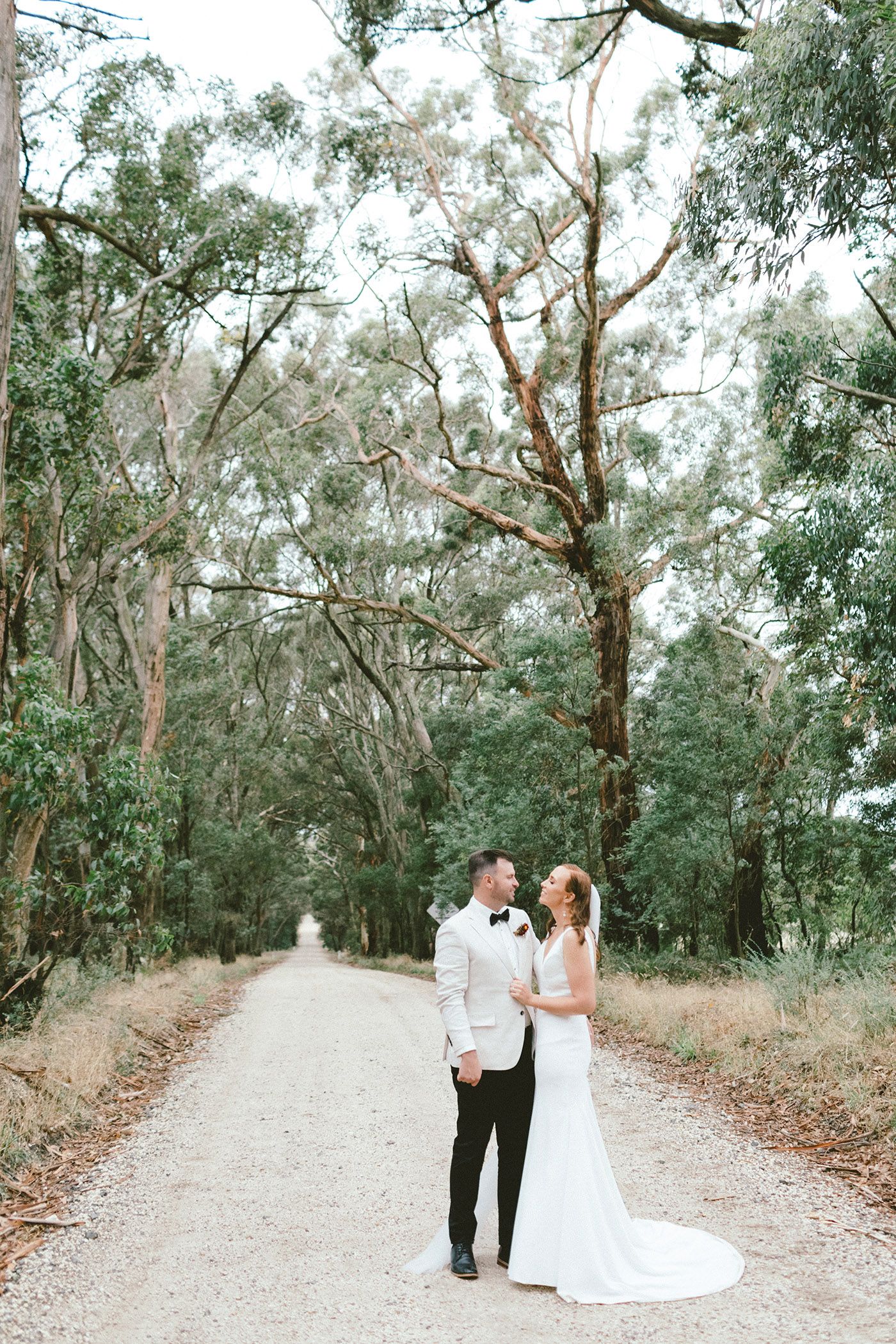 A couple standing on a tree-lined path in wedding attire, surrounded by eucalyptus trees.