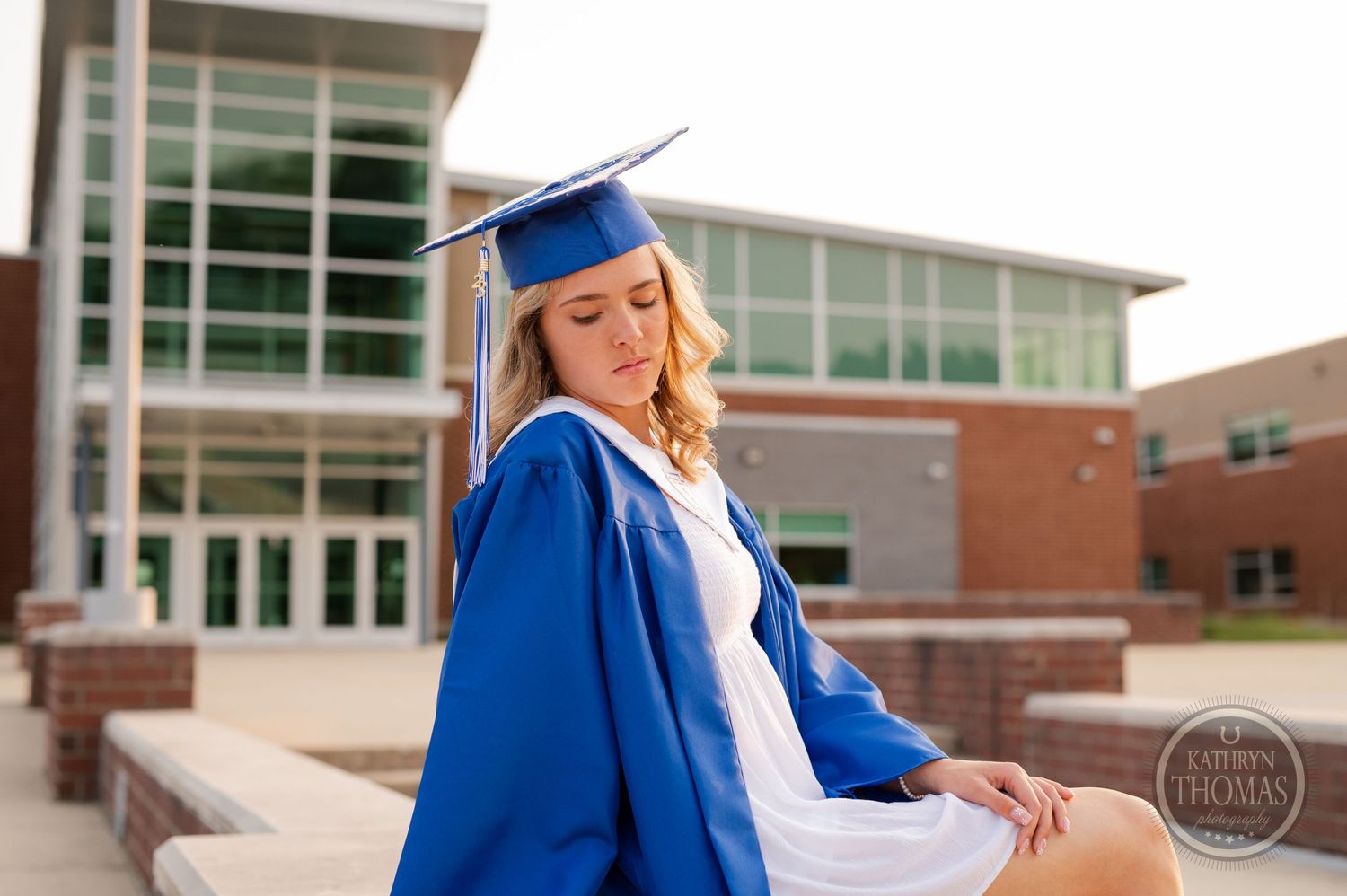 Graduate in blue cap and gown posing on steps outside modern school building.