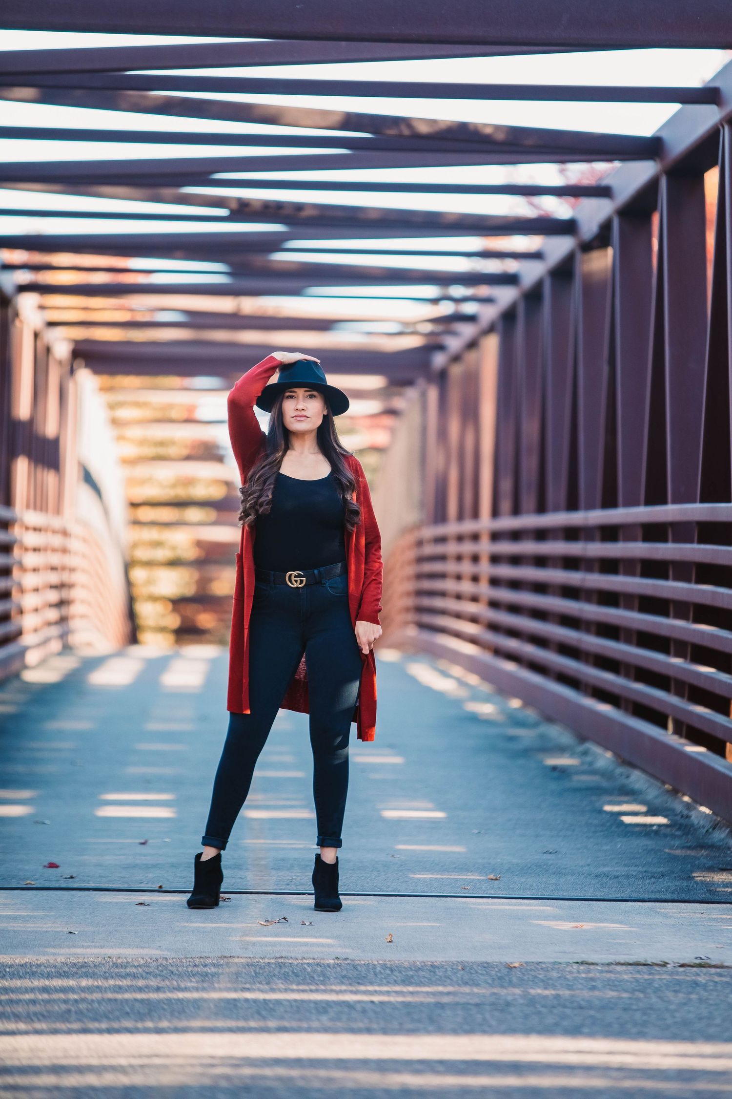 A fashionable woman in a red cardigan and black hat poses on a metal bridge walkway.