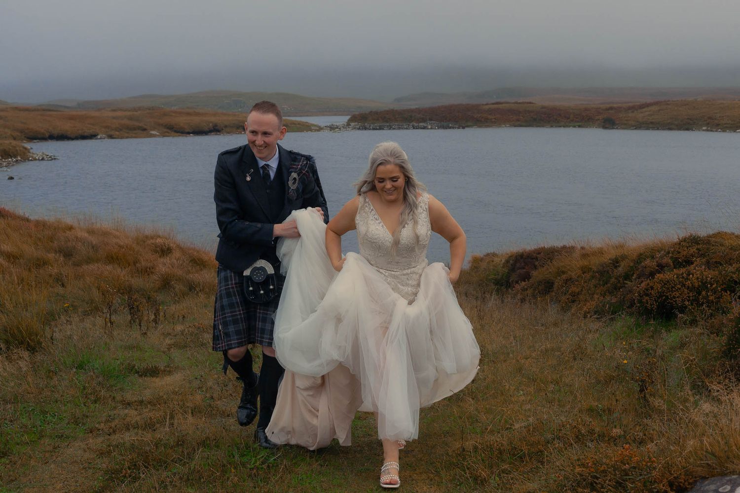 Groom helping bride with dress on muddy path.