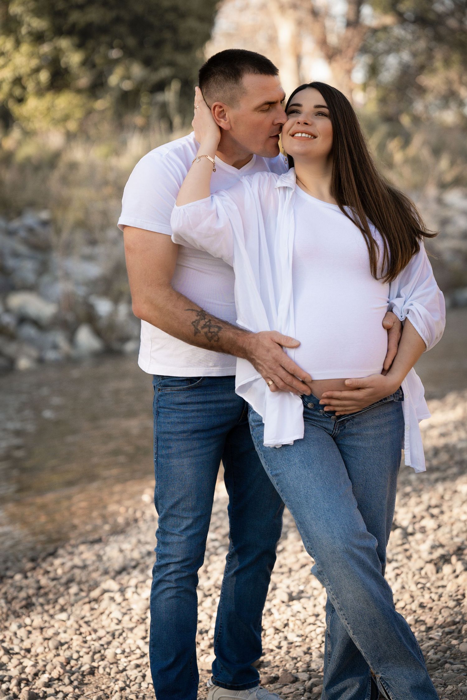 A romantic maternity photo of a couple in a sunlit forest, celebrating the upcoming arrival of their baby.