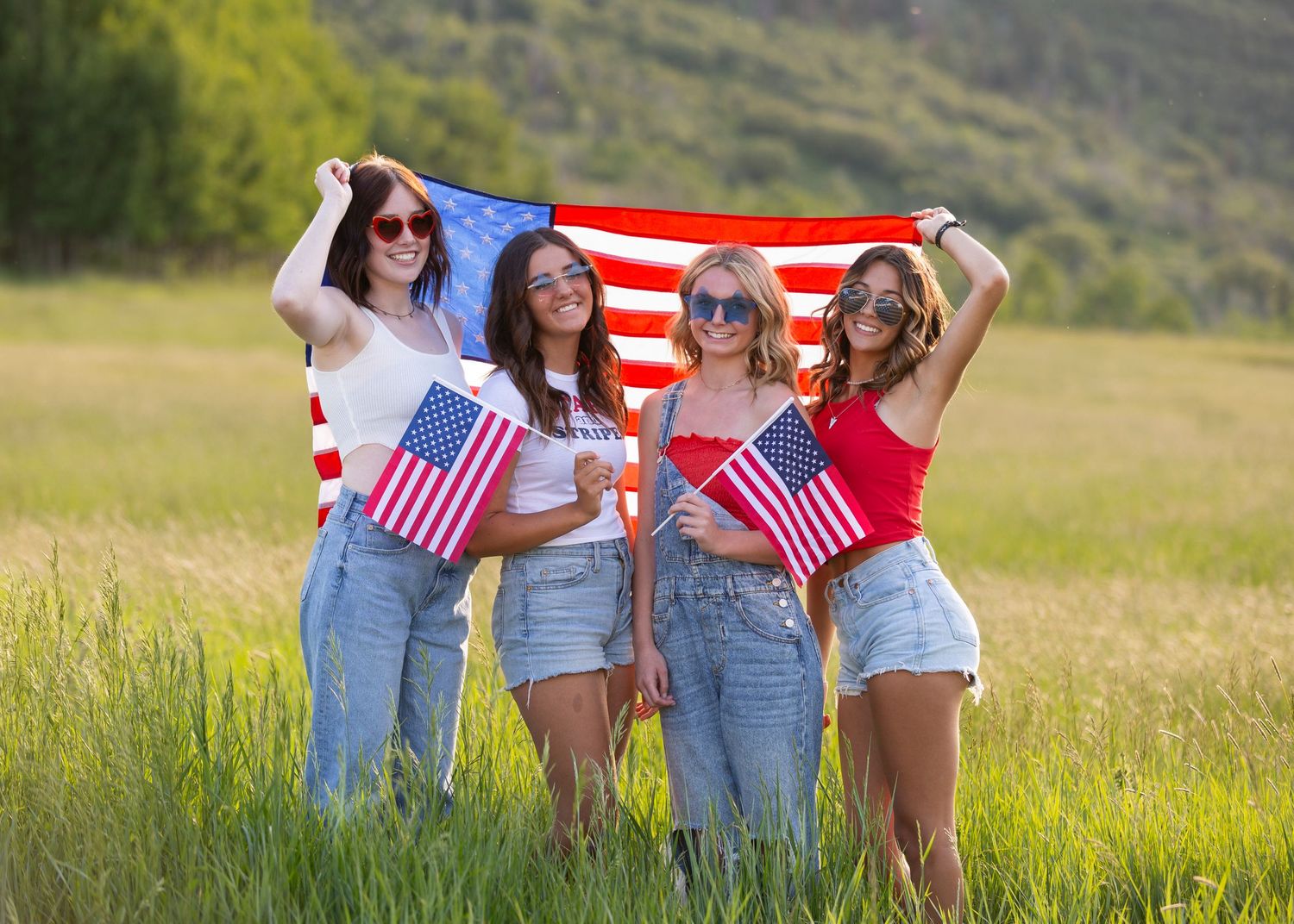 four senior girls in front of American flag with patriotic clothing and sunglasses on in a field