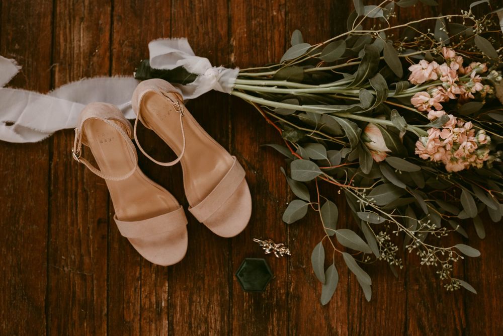 Nude bridal sandals and delicate floral bouquet arranged on rustic wooden surface.