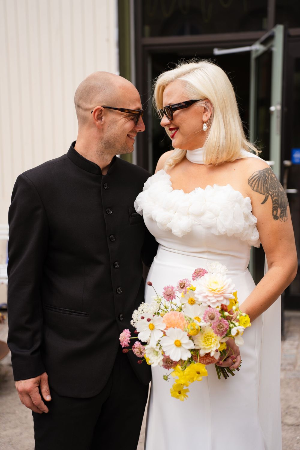 Wedding couple with colorful bouquet standing together against white building exterior.