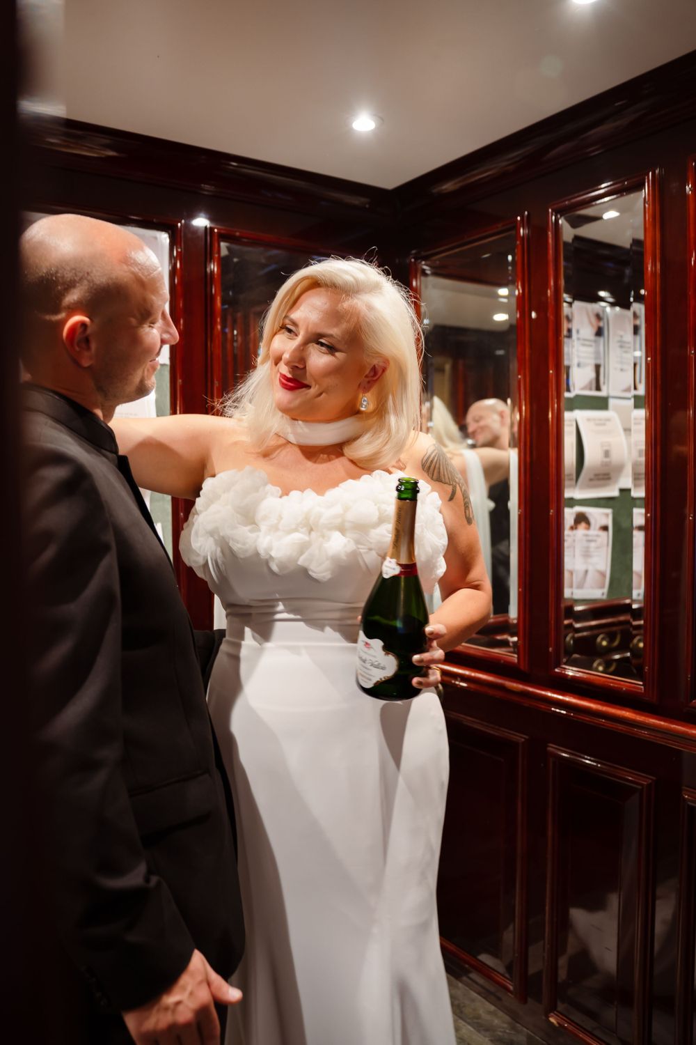 Elegant couple shares champagne moment by mirrored wall in formal attire.