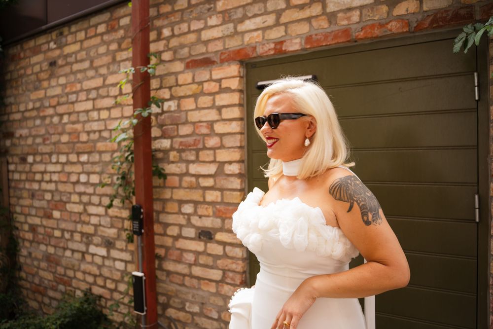 A bride in a white strapless gown and sunglasses poses against a brick wall on their wedding day.