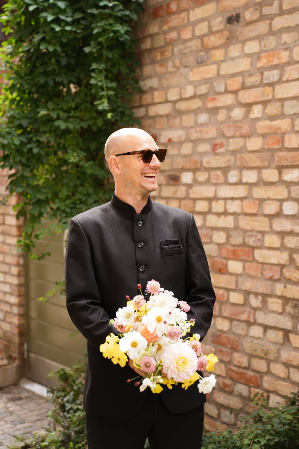 A person in a black mandarin collar suit holds a vibrant bouquet of pink and yellow flowers against a brick wall backdrop.