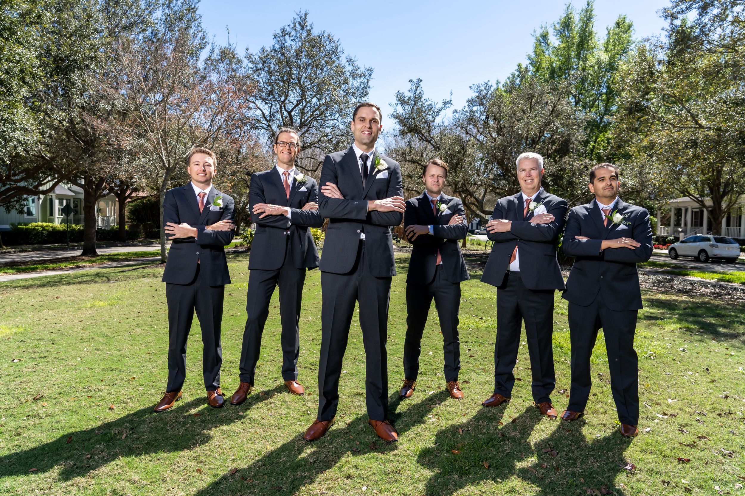 Groomsmen in black suits pose for formal wedding photo outdoors.