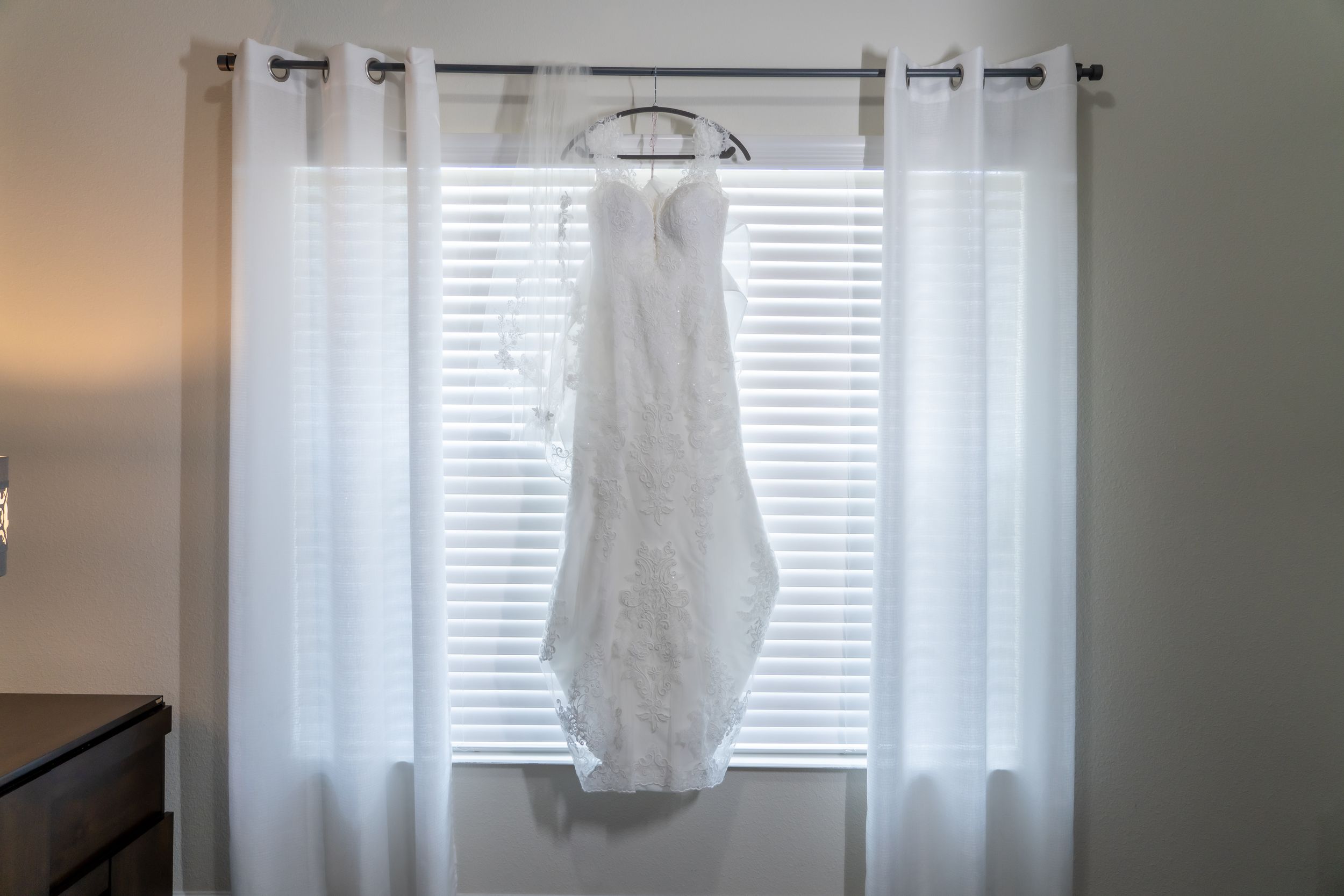 A white lace wedding dress hangs elegantly against a window with sheer curtains.