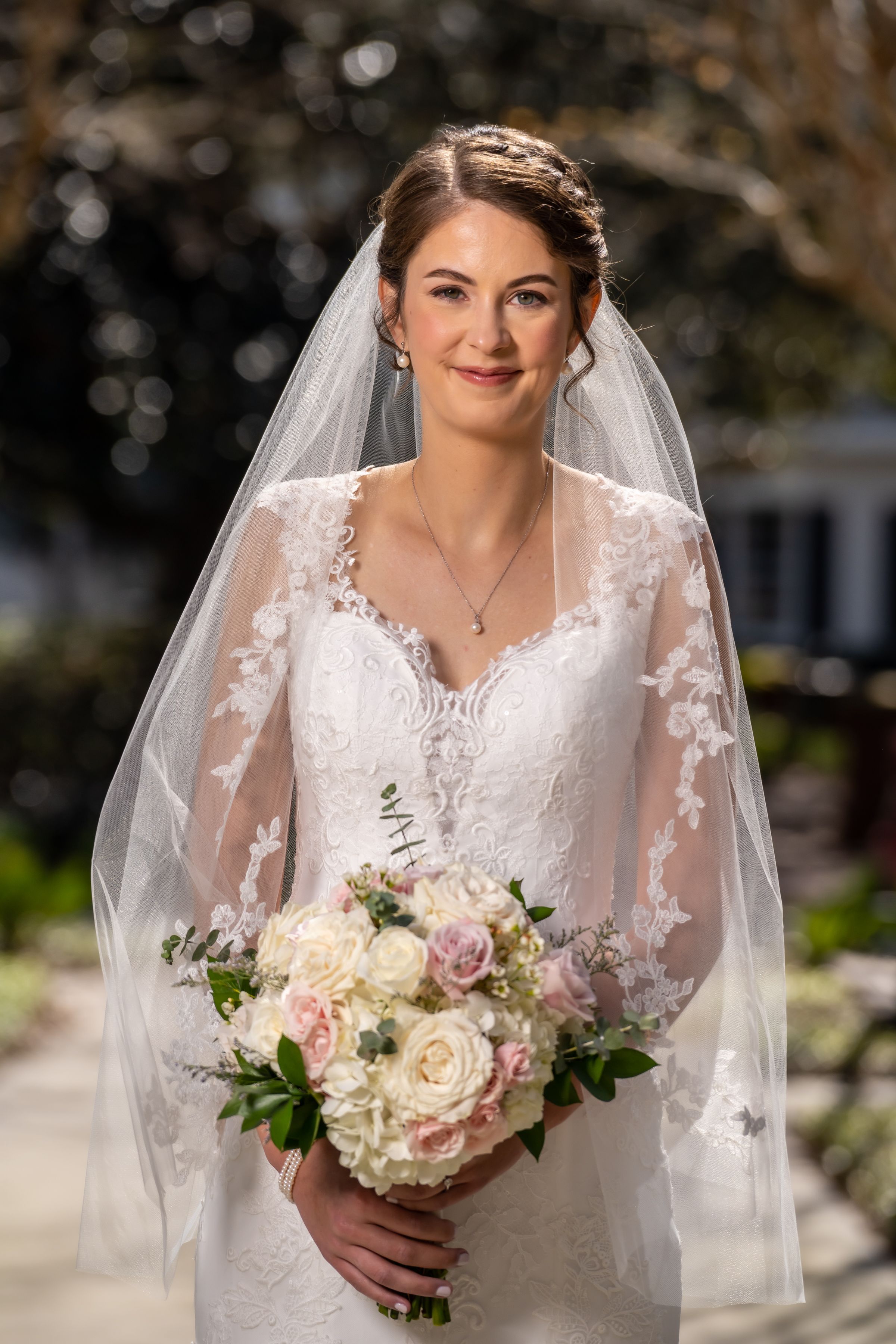 A bride in a white lace wedding dress with veil holds a bouquet of blush and ivory roses.